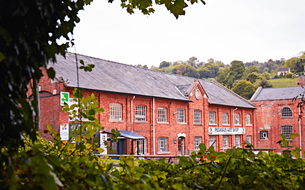 Pegasus Art at Griffin Mill, Thrupp, Stroud, Gloustershire. Large red brick mill building front of green trees.