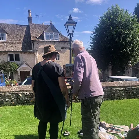 Roger Dellar guiding student painting on easel on lawn near stone cottage