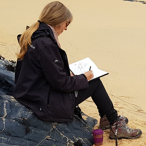 wendy rhodes drawing while sitting on a rock on the beach